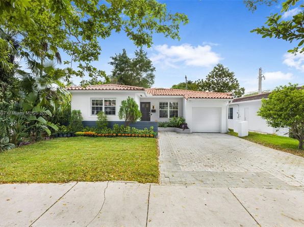 Front view of a single‑story Coconut Grove home with a red‑tile roof, paved driveway, and manicured lawn framed by tropical trees.
