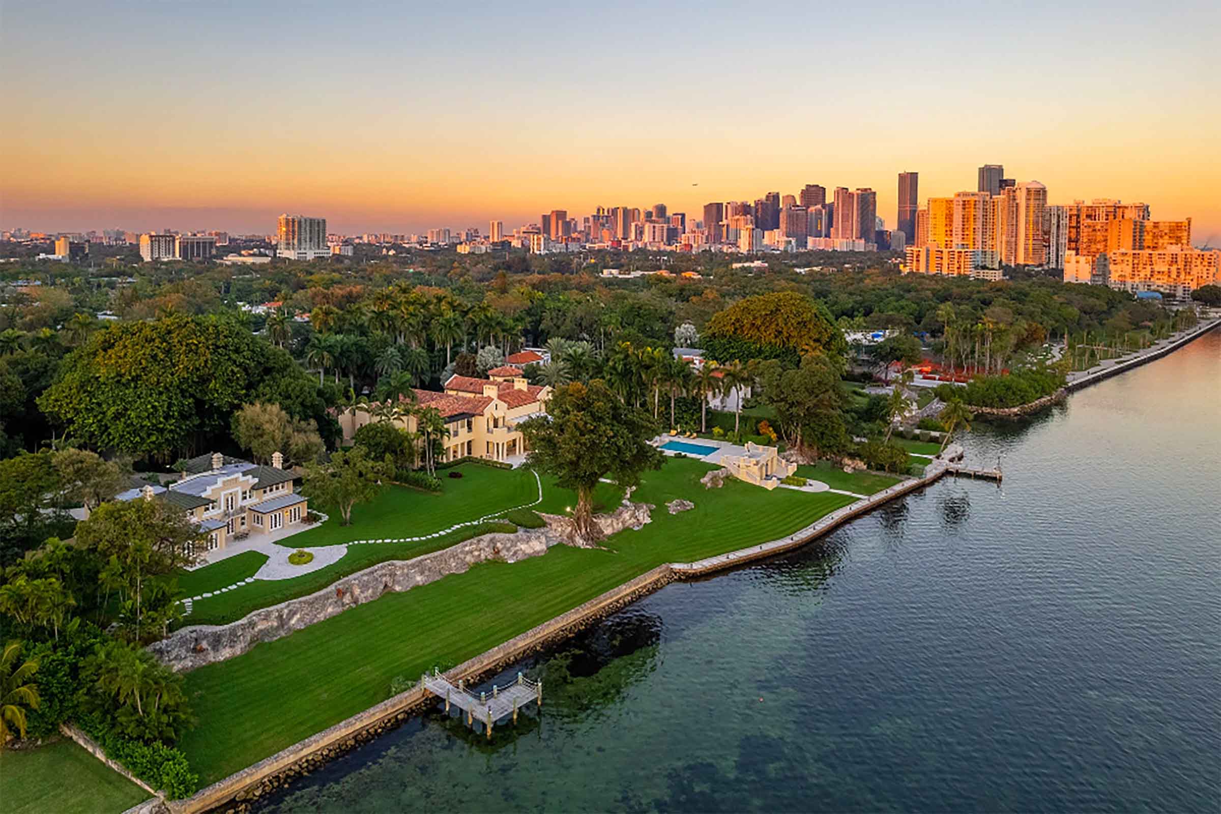 Aerial sunset view of waterfront Miami‑Dade estate with manicured lawn, private dock, and Coconut Grove skyline in the distance.