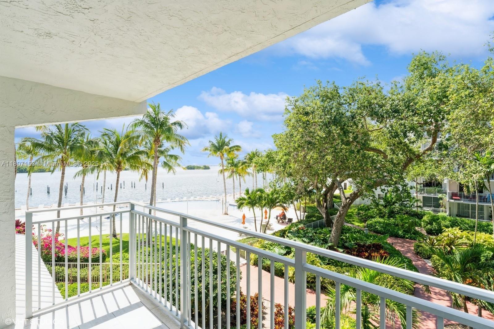 View from a balcony overlooking tropical landscaping, palm trees, and waterfront beach area with blue skies in a coastal Florida property