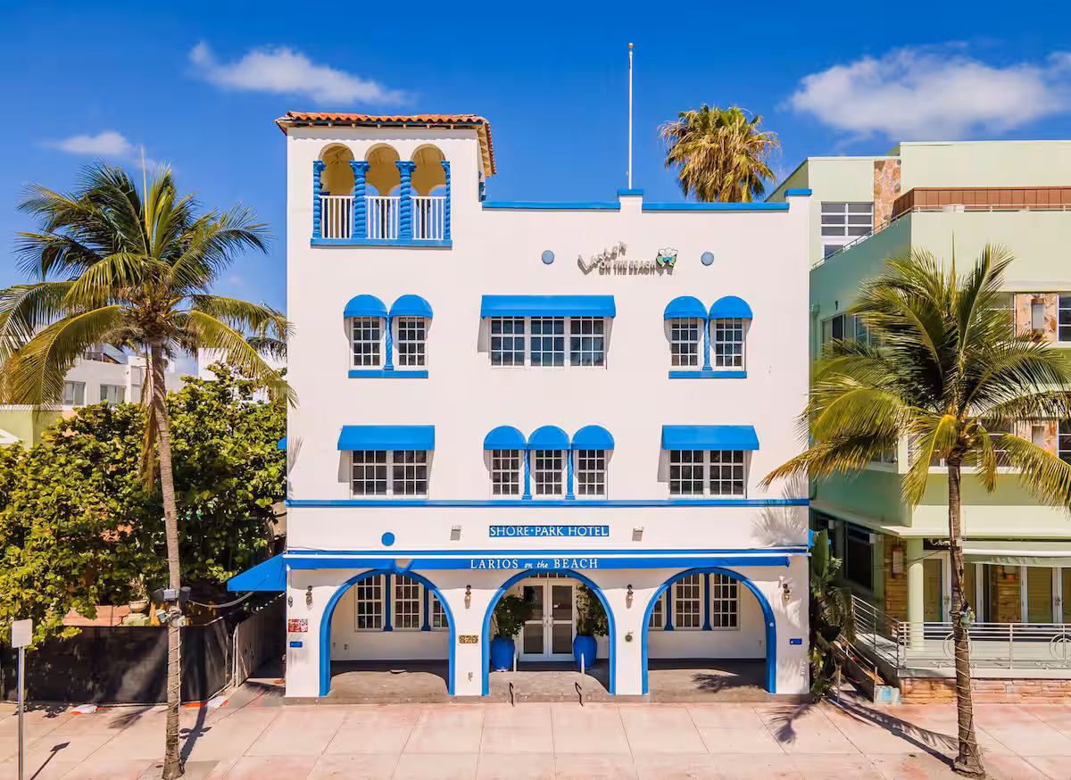 Historic Art Deco hotel with blue awnings in Miami Beach, framed by palm trees and a sunny sidewalk.