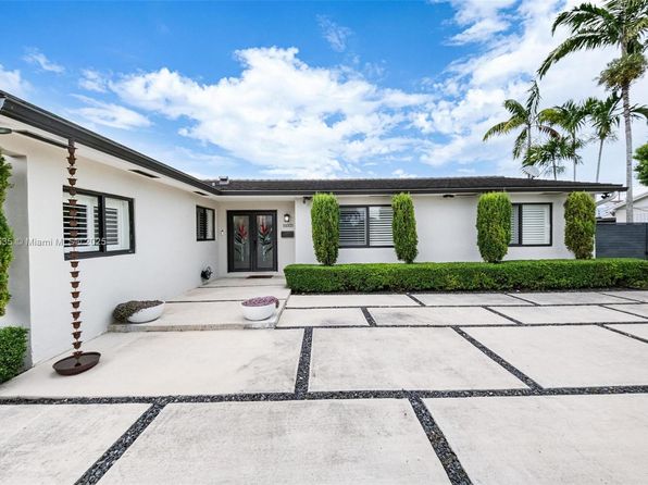 Modern white single-story home with black trim, palm trees, and a geometric paver driveway in South Florida