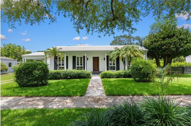 Front view of a traditional one-story home in Miami Shores with manicured lawn, palm trees, and symmetrical hedges under a blue sky